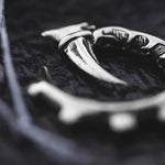 Close-up of the metallic, curved GIGER Biomechanical Open Ended Hoop Ear Weights in Silver by Kaya Movement, featuring intricate engravings against a textured background. The object, resembling biomechanical dangle hoop ear weights, appears to be part of a larger item and has a hook or claw-like shape. The dark, slightly out-of-focus background further highlights its details.
