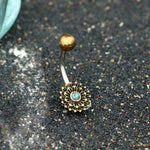 The MADA Lotus Flower Belly Piercing Ring in Gold & Turquoise by Kaya Movement features an exquisite bronze sunflower design with a small turquoise stone center, set against a textured dark gray background. Expertly crafted from surgical steel, the ring has a gentle bend and is adorned with a plain gold top ball. This unique piece of navel jewelry adds elegance to any look.