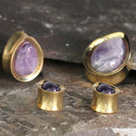 A set of exquisite jewelry featuring Kaya Movement's Amethyst Teardrop Stone Saddle Plugs in Gold, available in gauges from 12mm to 25mm, paired with smaller rounded purple gemstones also set in gold. The pieces are elegantly displayed on a textured dark surface.