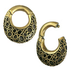 The SHAL Hoop Ear Weights in Gold with Clicker by Kaya Movement are designed as brass hoop ear weights with a 4 gauge. Featuring a textured pattern reminiscent of scales or stones and an antique gold finish, these earrings are completed with a secure clicker clasp fastening, adding a touch of vintage elegance to your piercing body jewelry collection.