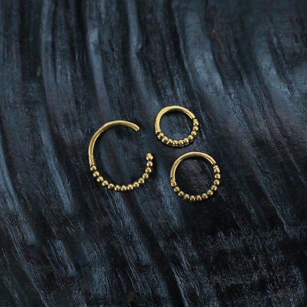 Two ORA Beaded Nose Rings in Gold (16 gauge) and one larger ORA Beaded Nose Ring in Gold (16 gauge) by Kaya Movement are displayed against a dark, textured background. The intricate beadwork adds detail and elegance to these beautiful nose rings inspired by tribal ethnic design.