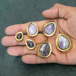A hand is holding six Amethyst Teardrop Stone Saddle Plugs in Gold from Kaya Movement, with gauges ranging from 12mm to 25mm. The gemstones are set in gold frames and vary in size, with the smallest on the left and the largest on the right, against a dark, textured background.