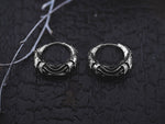 A pair of Silver Futuristic Hoop Earrings from Kaya Movement, inspired by cyberpunk aesthetics and ideal for tech lovers, rest on a textured black surface. Next to the earrings, thin dried twigs add an organic element to the dark, moody background.