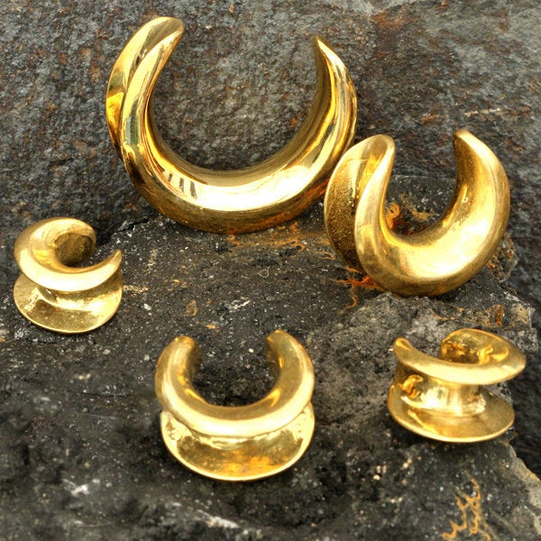 A set of Minimalist Horseshoe Saddle Tunnels in Gold by Kaya Movement, ranging from 4mm to 30mm gauge, are displayed on a textured, dark gray stone surface. The polished brass finish reflects light beautifully.