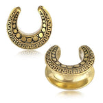 The DAG Tribal Horseshoe Saddle Tunnels in Gold by Kaya Movement feature a pair of gold-colored steel saddles adorned with intricate black and gold detailing, including small circular patterns and wavy lines along the edge. The curved, horseshoe-shaped design is open at the top and showcases a reflective, polished finish. Available in gauges ranging from 8mm to 25mm.