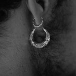 A close-up shot of an ear adorned with two distinctive earrings. The upper earring is a small, textured hoop, while the lower piece, the AARO Cyborg Clicker Hoop Septum Nose Ring in Silver from Kaya Movement, features a larger, intricate hoop with a segmented design that evokes dystopian body jewelry. Both pieces add a futuristic edge to the monochromatic image.