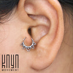 Close-up of an ear adorned with a decorative hoop earring featuring an ornate, triangular pattern. Crafted from 925 Sterling Silver, the intricately designed KAAL Lotus Ring in Silver by Kaya Movement adds elegance. The words "Kaya Movement" are visible in the bottom left corner of the image.