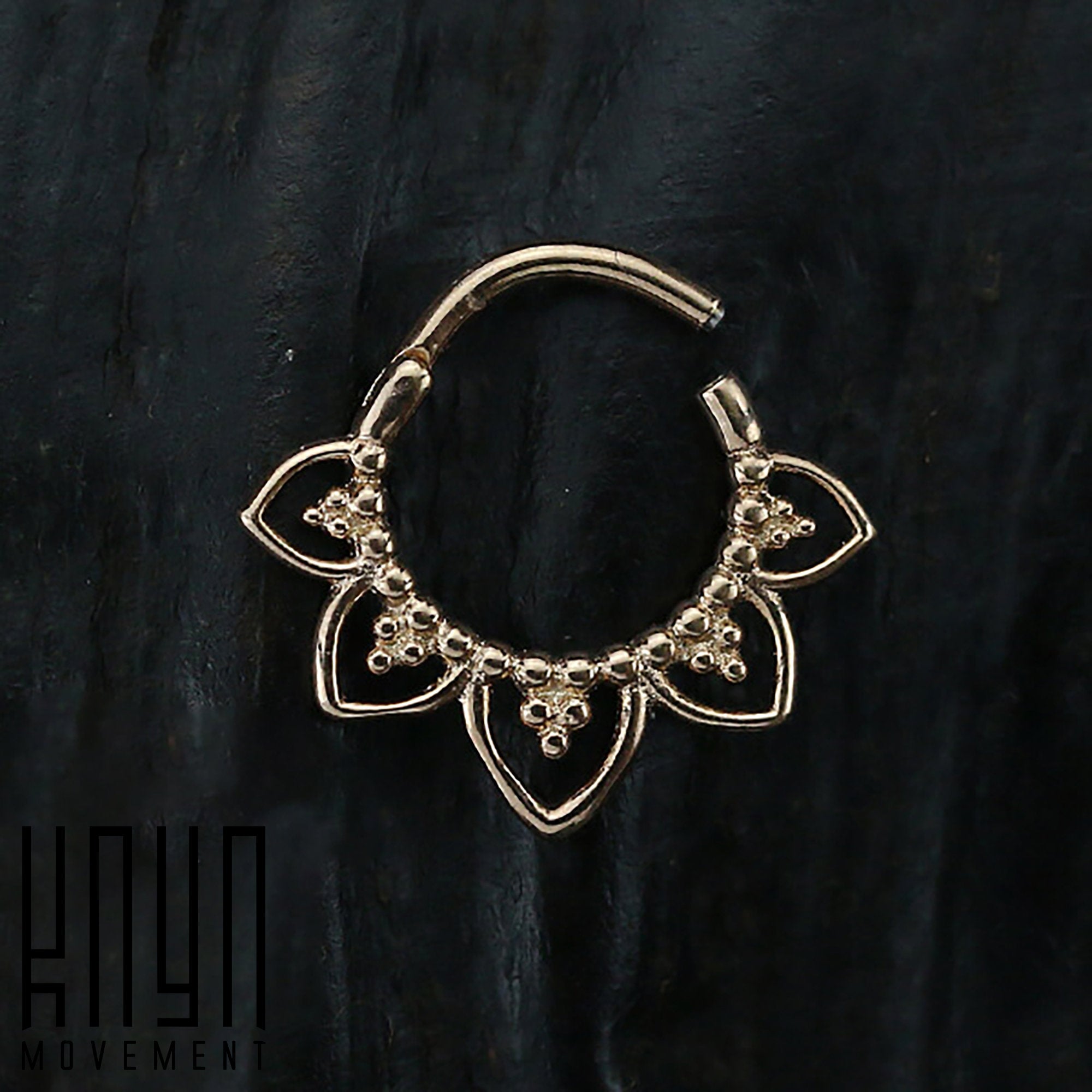 The MAWAR Lotus Clicker Septum Ring in Rose Gold, available in a 16 gauge size, is exhibited against a dark, textured backdrop. This gold-hued ring showcases an intricate flower-like design with small, delicate loops and bead-like details on the lower portion. Crafted from surgical steel with a rose gold PVD coating, the "Kaya Movement" logo is visible at the bottom left corner of the image.