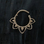 The MAWAR Lotus Clicker Septum Ring in Rose Gold, available in a 16 gauge size, is exhibited against a dark, textured backdrop. This gold-hued ring showcases an intricate flower-like design with small, delicate loops and bead-like details on the lower portion. Crafted from surgical steel with a rose gold PVD coating, the "Kaya Movement" logo is visible at the bottom left corner of the image.