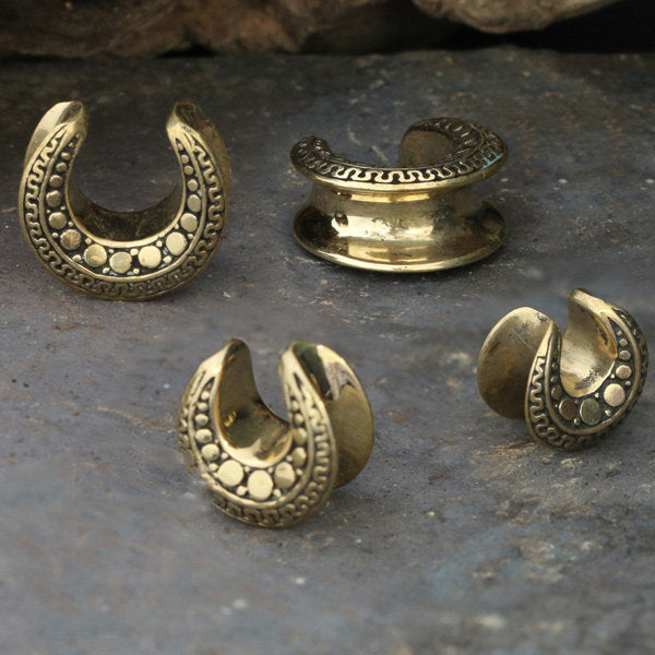 A set of DAG Tribal Horseshoe Saddle Tunnels in Gold by Kaya Movement, available in 8mm to 25mm gauge, are displayed on a stone surface. The jewelry pieces feature intricate etched patterns and dot designs, with some shaped like horseshoes and others forming complete circular bands.