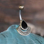 The Kaya Movement IKOSA Fan Belly Piercing in Gold & Black Mother of Pearl, featuring a gold-colored charm with an intricately designed teardrop-shaped black mother of pearl stone, is displayed against a blurred, blue-toned background. This 14 gauge navel ring includes a curved surgical steel barbell with a small gold ball at the top.