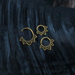 A close-up photo of two tiny Kaya Movement ILA Seamless Septum Rings in Gold (16 gauge) with ornate petal-like designs, displayed on a dark, textured background. The intricate patterns on the septum rings resemble small lotus flowers and highlight the metallic shine against the contrasting dark surface.