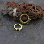 A pair of SPIKE Punk Spike Nose Rings in Gold by Kaya Movement rests on a textured surface of rough, brownish rock. The rocks have an irregular, porous structure, providing a rustic backdrop to the metallic rings. Reminiscent of gold brass body jewelry, one ring is slightly larger and both display a mechanical, spiked design.