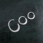 Three silver hoop earrings of different sizes are arranged in a row on textured black fabric. The largest hoop is incomplete with an intentional gap, followed by two smaller, complete hoops. Crafted from 925 Sterling Silver and designed in a minimalist style by Kaya Movement, the PLATO Seamless Ring in Silver | 16 gauge is showcased prominently with the font "krny" appearing in the bottom left corner of the image.