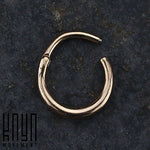 A close-up of a small, circular rose gold AMAO Clicker Nose Ring in 316L Surgical Steel, placed on a dark, textured surface. The nose ring appears to have a hinge or clasp mechanism. The logo "Kaya Movement" is positioned in the bottom left corner of the image.