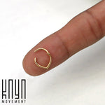 A close-up of a person's finger displaying a small, gold body jewelry ring with a minimalist design, balanced on the fingertip. The teardrop-shaped ring is crafted from surgical steel and part of the TIKA Teardrop Septum Clicker Ring in Gold collection by Kaya Movement. The logo "Kaya Movement" is visible in the bottom left corner.