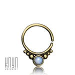 The ANI Seamless Septum Ring in Gold & Moonstone, a 16 gauge piece adorned with a shimmering blue moonstone and small decorative beads around the base, is showcased against a white background. In the bottom left corner, the brand logo "Kaya Movement" emphasizes this elegant piece of body jewelry.