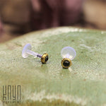 A pair of KEA Labret Studs in Gold & Onyx (16 gauge) lies on a smooth, green, shiny surface. Beside them, the words "Kaya Movement" are subtly visible in the bottom left corner. The background is blurred, emphasizing the minimalist stud earrings crafted with Black Onyx stone jewelry in the foreground.