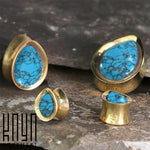 A gold jewelry set featuring blue turquoise stones with black veining, including two rings and two brass teardrop saddle plugs. Each piece boasts teardrop-shaped turquoise stones and is elegantly displayed on a stone surface with the "Kaya Movement" logo in the corner.