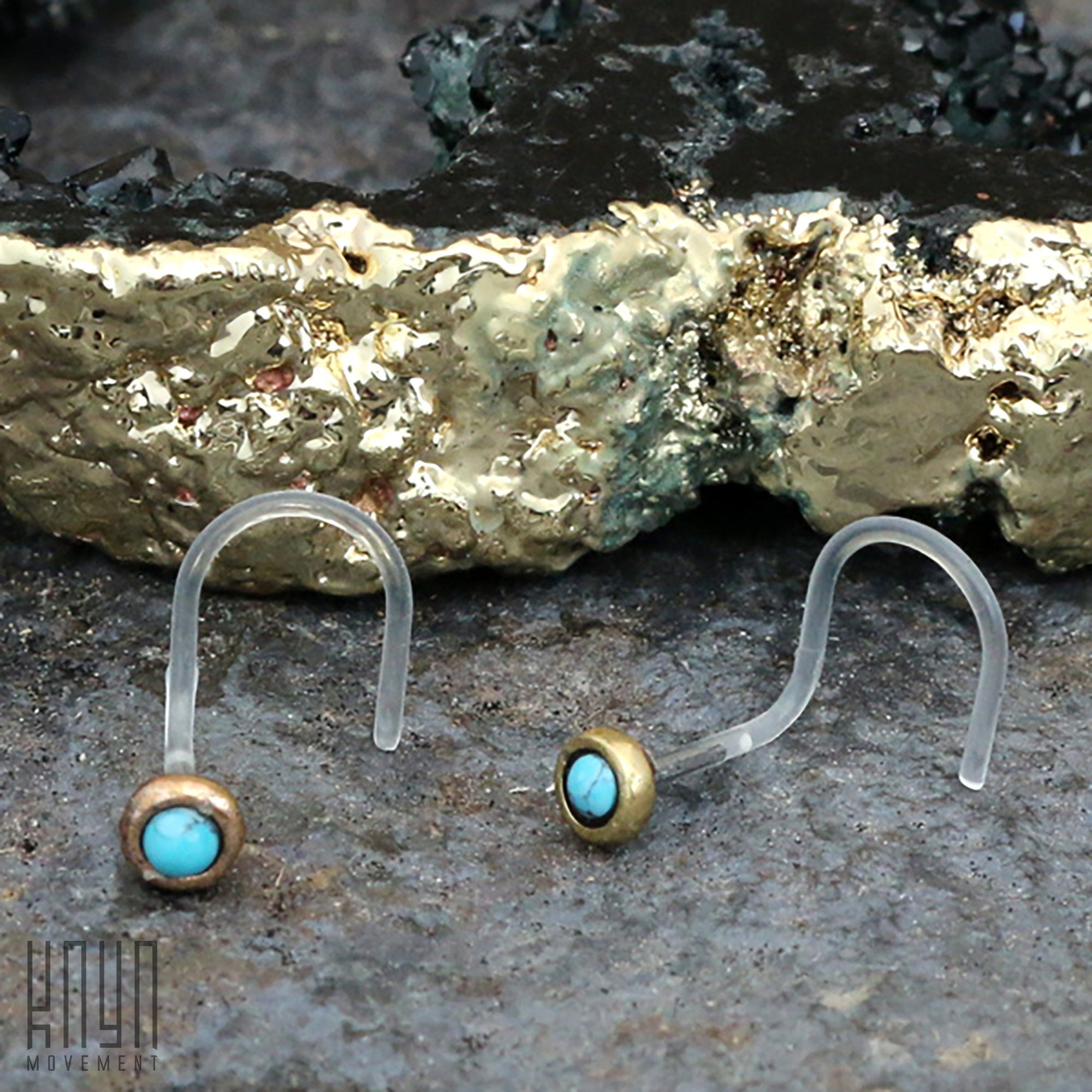 The EDEN Nose Screw Studs in Gold with Turquoise from Kaya Movement are displayed as a pair. These 20-gauge nose screws feature transparent bio-plastic hooks and round, flat settings that securely hold natural Turquoise stones encased in metallic bezels. The studs are placed on a rough, textured surface with a gold and black backdrop.