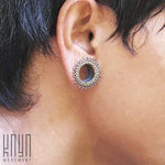 A close-up of a person's ear showcases the intricate, circular ornamentation of the TIMO Sacred Lotus Mandala Round Tunnels in Silver from Kaya Movement. The earring features a dark center with detailed edges that evoke the charm of a handcrafted vintage piece. The person sports short, dark hair, and the "Kaya Movement" logo is visible in the corner.