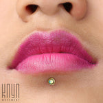 Close-up of a person’s lips adorned with bright pink and purple lipstick. A small, shiny piercing featuring Kaya Movement's KEA Labret Stud in Gold & White Opal (16 gauge) is visible just below the lower lip. The word "Kaya Movement" is visible in the bottom-left corner of the image.