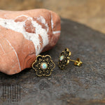 A pair of Opal Studs Earrings - Boho Piercing with floral designs and opal centers are placed on a stone surface. One earring is leaning against a textured reddish rock. The background is blurred, featuring earthy tones. The logo "Kaya Movement" is visible in the lower-left corner.