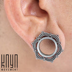 A close-up image of an ear showcasing a handcrafted LOTUS Sacred Lotus Mandala Round Tunnel in Silver, designed with a floral pattern and a hollow center. The plug is seated in the lobe piercing, and the "Kaya Movement" logo is placed in the bottom left corner of the image.