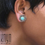 Close-up of a person wearing Lotus Flower Saddle Plugs in Gold & Turquoise, featuring a turquoise stone at the center. The person's short, dark hair partially covers their ear. "Kaya Movement" is written in black text at the bottom left corner of the image.