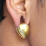 Close-up of a person's ear adorned with the remarkable Kaya Movement OLO Minimalist Teardrop Ear Hangers in Gold. These 14mm gauge earrings boast a sleek, polished finish and an unconventional design that forms a thick, teardrop shape, hugging the earlobe closely to create a bold, contemporary look.