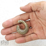 A hand holding a small, round piece of body jewelry gauge earring with an intricate flower of life pattern. The metallic jewelry, the FLOWER OF LIFE Hoop Ear Weights in Gold with Clicker from Kaya Movement, appears to be a pendant or an earring. The text "Kaya Movement" is visible in the bottom left corner of the image.