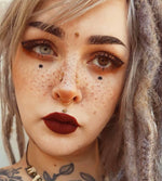 A close-up of a person with light brown eyes, freckles, and heavy makeup, including winged eyeliner and dark red lipstick. They have multiple facial piercings adorned with Kaya Movement's LUST Flower Labret Stud in Gold (16 gauge), bleached dreadlocks with dark roots, and tattoos visible on their neck.