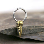 A small, intricately designed keychain featuring Kaya Movement's CROW SKULL Gothic Silver Ring, available in 18, 16, or 14 gauge, adorned with a gold charm and attached to a circular metal ring. The keychain rests on a dark, textured stone surface. The bird skull charm boasts a detailed beak and hollow eye sockets, offering a realistic appearance.