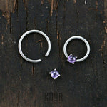 A small circular Kaya Movement ORASA Nose Ring in Silver & Amethyst (16 gauge) along with an open silver nose ring and a matching purple gemstone earring stud are arranged on a dark wooden background. The image, showcasing exquisite silver body jewelry, has a textured, rustic feel.