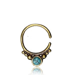 The ANI Seamless Septum Ring in Gold & Turquoise by Kaya Movement is a 16 gauge piece of body jewelry with an antique-style circular design. It features a central turquoise gemstone and small decorative brass spheres symmetrically arranged around the bottom, beautifully displayed against a white background.