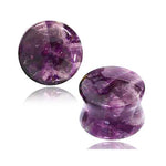 Two small polished stones, the Amethyst Round Stone Saddle Plugs by Kaya Movement, both featuring a deep purple color with lighter streaks. One stone is disc-shaped, and the other has a convex form reminiscent of saddle plugs. They rest side by side on a white background, evoking the calming energy of the third eye.