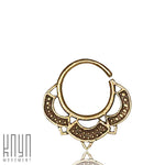 The MANDA Mandala Septum Ring in Gold by Kaya Movement is an 18-gauge, gold-colored ornate hoop nose ring featuring an intricate design at the bottom with small decorative cutouts and patterns. Showcasing the elegance of gold body jewelry, the ring is displayed against a plain white background with a logo in the bottom left corner that reads "Kaya Movement.