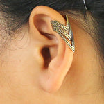 A close-up of an ear adorned with the IRA Chevron Ear Cuff in Gold by Kaya Movement. This unique gold-toned ear cuff showcases a geometric, triangular design with intricate detailing and a hint of tribal flair, elegantly wrapping around the upper part of the ear. The hair is pulled back to showcase the accessory.