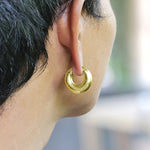 Close-up of a person's ear wearing the DAYA Minimalist Hoop Ear Weights in Gold with Clicker by Kaya Movement. The unisex body jewelry, available in 2 gauge, has a smooth, polished finish. The background is blurred, highlighting the earring. The person's hair is short and dark.