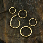 A collection of Kaya Movement's AMOI Clicker Nose Rings in Gold, available in 20, 18, 16, or 14 gauge sizes, is arranged on a textured dark brown background. The assortment includes rings ranging from small to medium sizes with both closed and partially open designs. This rustic setting accentuates their beauty and versatility for multi-use piercings.