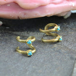 A close-up image of the LORY Ear Cuff in Gold & Turquoise by Kaya Movement shows two gold ear cuffs adorned with small blue turquoise stones, placed on a textured gray surface. The minimalist design is enhanced by a pink, shell-like object in the background, providing a striking contrast to the jewelry pieces.