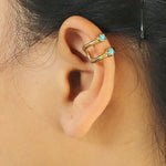 A close-up of a person's ear showcases the minimalist LORY Ear Cuff in Gold & Turquoise by Kaya Movement, adorned with two small blue turquoise gemstones. The elegant cuff is positioned around the upper part of the ear cartilage, while the person's hair is partially visible and tucked behind their ear.
