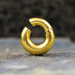 A close-up of the Kaya Movement Minimalist Steel Hoop Ear Weights in Gold with Magnet Closure (2 gauge) displayed upright on a stone surface. The background is blurred and dark, which highlights the ear weights' shiny, reflective finish, adding an understated elegance to their aesthetic.
