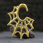 The Spider Web Gothic Steel Ear Hangers in gold by Kaya Movement, designed as a 2 gauge piece, rest on a dark, textured stone surface. The small golden hanger integrates an intricate spider web design and contrasts beautifully with the rough texture, evoking the precision and care reminiscent of tribal design ear weights.