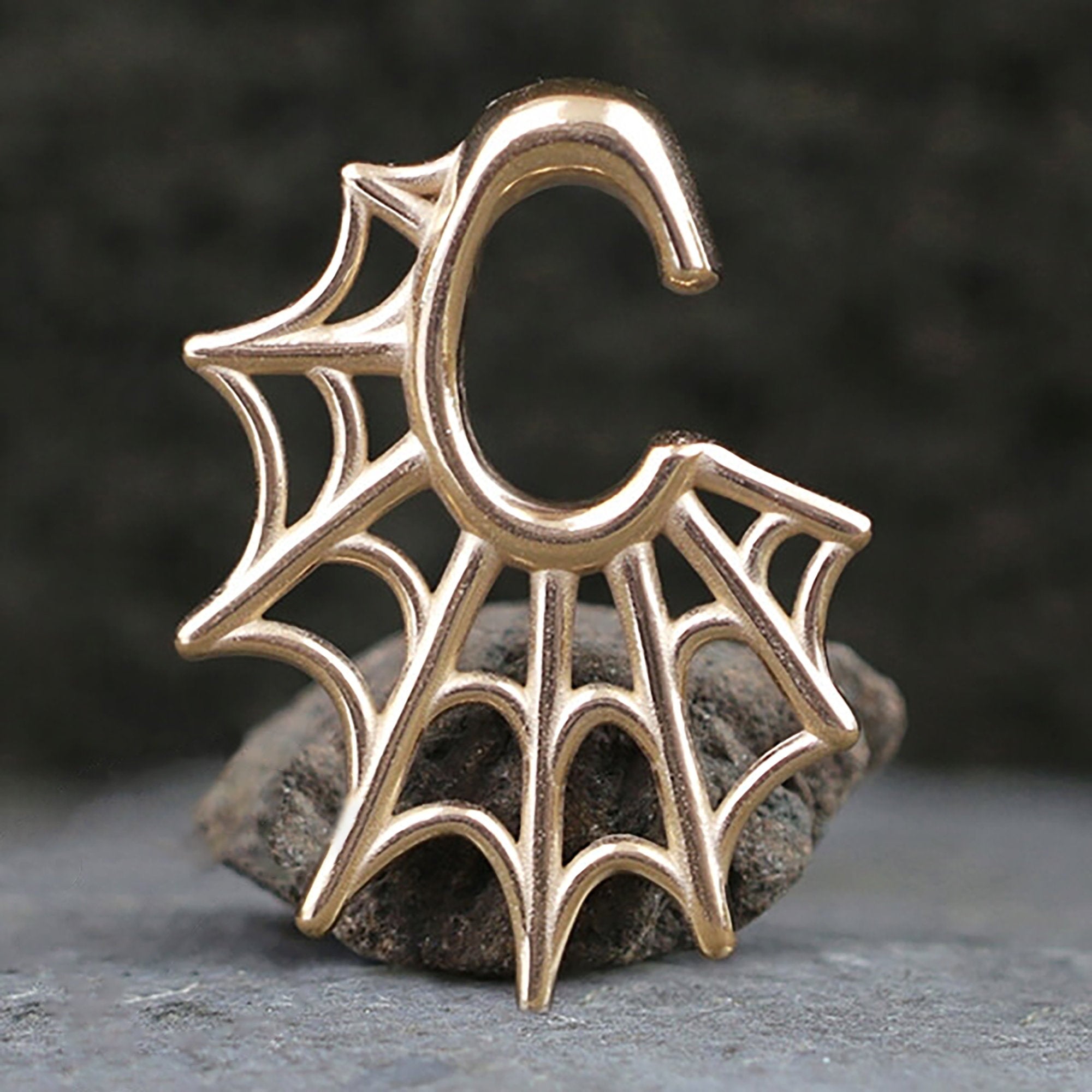A pair of Spider Web Gothic Steel Ear Hangers in Rose Gold by Kaya Movement, shaped like the letter "C" with an intricate spider web design reminiscent of tribal ear weights, rest on a stone with a blurred dark background.