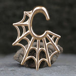 A pair of Spider Web Gothic Steel Ear Hangers in Rose Gold by Kaya Movement, shaped like the letter "C" with an intricate spider web design reminiscent of tribal ear weights, rest on a stone with a blurred dark background.