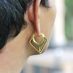A close-up of a person's ear adorned with Kaya Movement's Steel Triangle Ear Weights in Gold with a magnet closure, featuring a triangular shape and accented by spherical details and extending rays. The short hair accentuates the intricate pattern of these exquisite 2 gauge earrings.