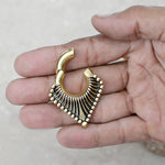 A hand holds the Kaya Movement Steel Triangle Ear Weights in Gold with Magnet Closure (2 gauge), showcasing an ornate, gold-plated PVD earring with a distinctive angular design. The earring displays a pattern of small golden beads and black detailing, forming a geometric, fan-like shape. The background is blurred and neutral.