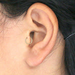 Close-up of a person's ear adorned with the AMOI Clicker Nose Ring in Gold from Kaya Movement, delicately placed in the earlobe. The person has dark hair, partly secured with a black pin or clip.