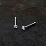 A pair of Kaya Movement Tiny Diamond Silver Nose Pin Studs (20g) with Zircon Crystal Gemstones, crafted from surgical steel 316L, are displayed on a dark, textured surface. The crystals are held by four-prong settings, and the studs' posts catch and reflect a bit of light.